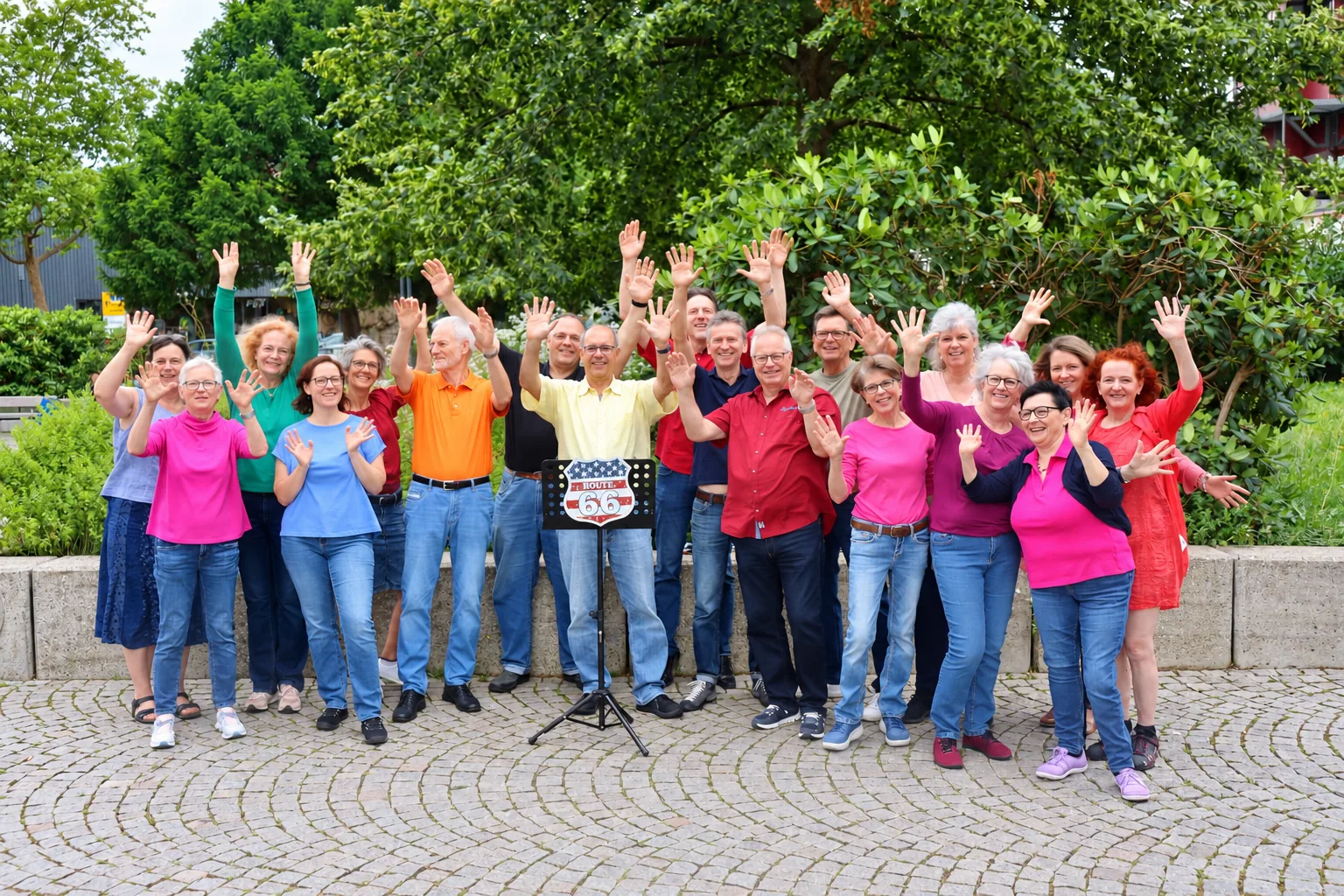 Mitglieder des Route66 Chors winken gemeinsam vor einer grünen Gartenkulisse.